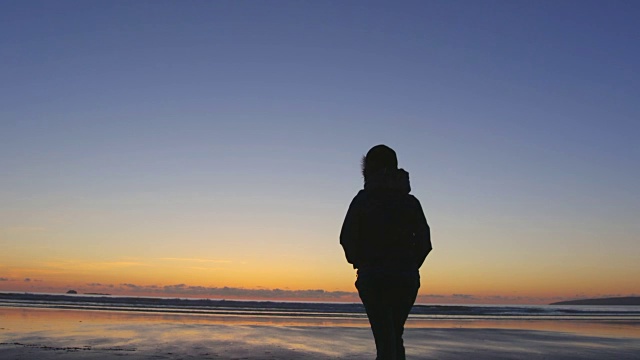 在日落时，在蓝天下的海滩上行走的剪影女人视频素材
