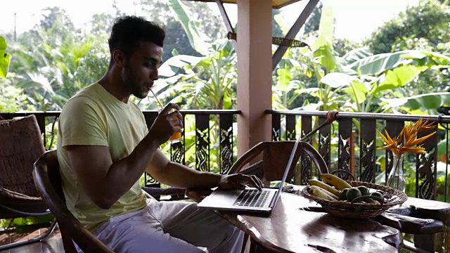 一个年轻人在笔记本电脑上打字，在阳台上吃早餐，混合种族，一个人一边工作一边喝果汁，坐在户外的桌子旁视频素材