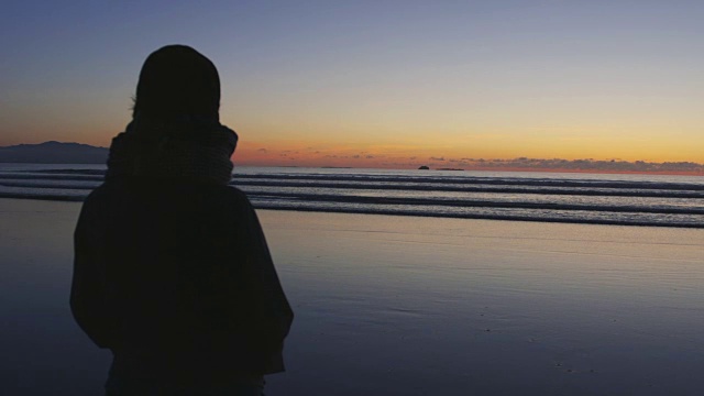 在日落时，穿着冬装站在海滩上的剪影女人视频素材