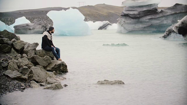 年轻英俊的男子坐在冰岛jokul沙龙冰泻湖的岩石上欣赏冰川的景色视频素材