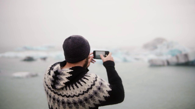 年轻时尚男子站在冰岛Jokulsalon冰礁湖的岸边，用智能手机拍照视频素材