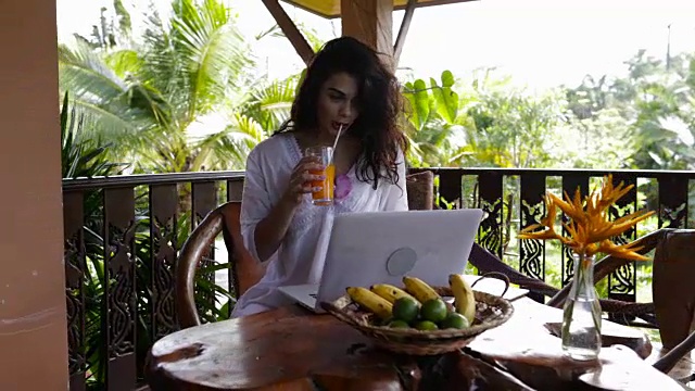 年轻女孩在夏天的阳台上用笔记本电脑吃早餐，美丽的女人一边喝果汁一边打字视频素材