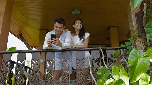 年轻夫妇在阳台上仰望交谈的男人和女人拥抱拥抱使用手机幸福的微笑视频素材