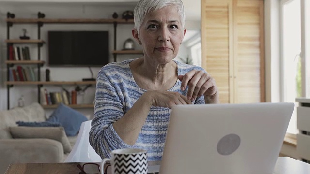 快乐成熟的女人在家里使用笔记本电脑，并在看相机时竖起大拇指。视频素材