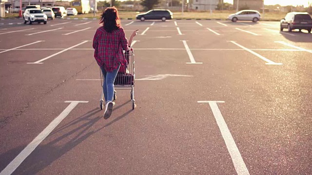 一个年轻女人坐在购物车里，而她的朋友在购物中心的停车场推着她，享受户外购物手推车比赛在日落视频素材