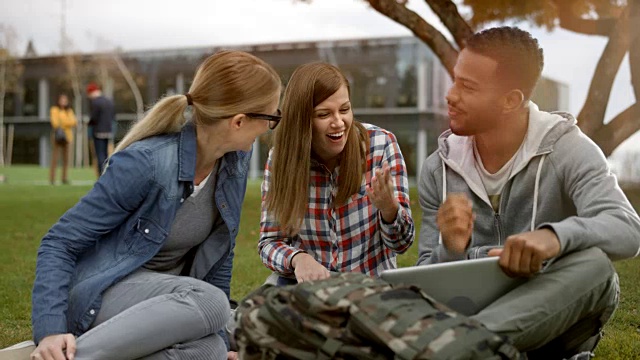 两个白人女学生坐在公园里，他们的多种族男性朋友拿着一台笔记本电脑视频素材