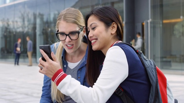 亚洲女学生和她的白人女性朋友一边笑一边聊天一边刷手机视频素材