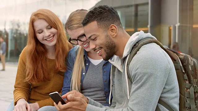 两名白人女学生和一名多民族青年男子在学生大楼前查看智能手机视频素材