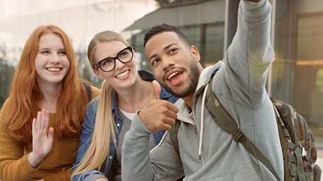 多种族的男学生和他的两个白人女性朋友在学生大楼前用智能手机拍视频视频素材