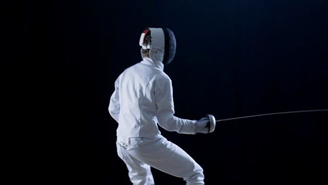 侧面的一个装备齐全的熟练击剑运动员训练他的攻击和刺与陪剑。拍摄孤立的黑色背景。视频素材