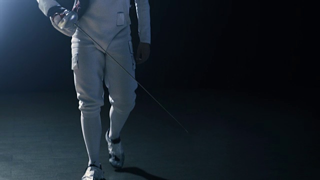 英俊的年轻击剑手走进聚光灯戴上防护面具举起剑准备比赛。拍摄孤立的黑色背景。视频素材