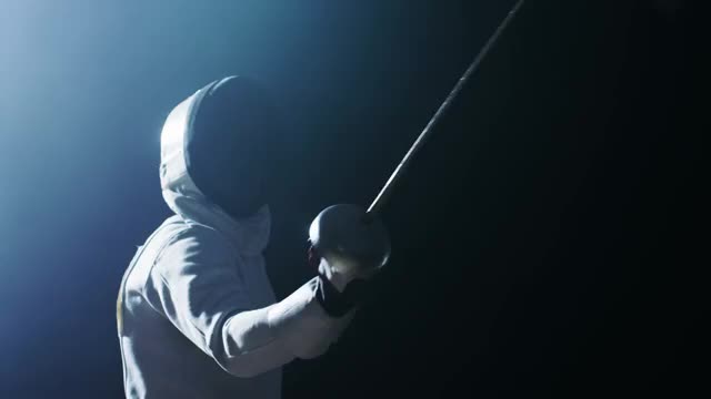 装备齐全的熟练击剑运动员训练用花剑。他在聚光灯下，周围是黑暗。他练习攻击，防御，跳跃，刺和弓步。拍摄孤立的黑色背景。视频素材