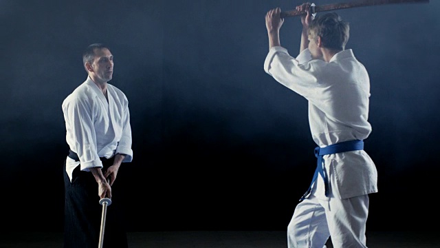 武术大师教他的年轻学生如何与木剑Bokken战斗。对练在几秒内结束，主人获胜。拍摄是孤立的黑色背景和慢动作。视频下载