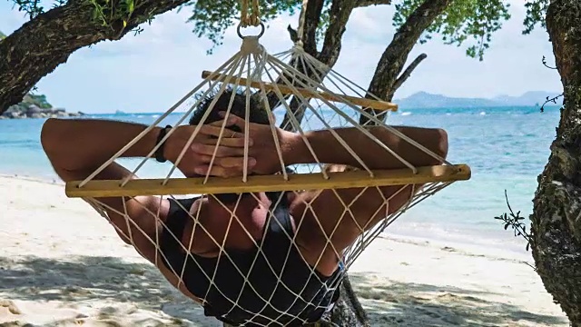 在美丽的蓝色海洋和其他岛屿的背景前，一个男人在沙滩上的吊床上放松地摇摆。在树荫下躲避阳光。视频下载