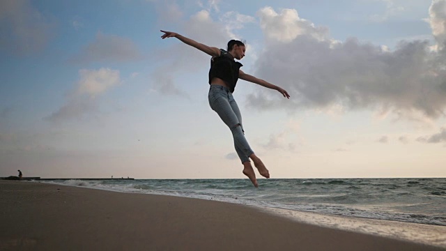 年轻女子穿着休闲风格-牛仔裤和黑色上衣在海滩上跳芭蕾。迷人的芭蕾舞演员在秋季的海岸线上练习跳跃。慢动作视频素材