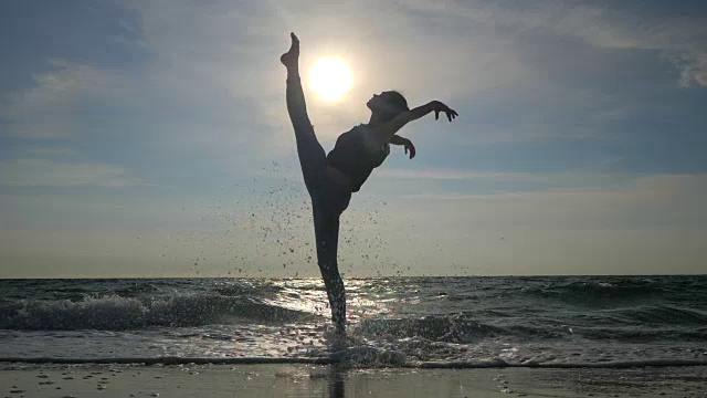 剪影的年轻女子在休闲风格-牛仔和黑色上装做芭蕾在海滩上。迷人的芭蕾舞演员在秋天的沙滩上练习伸展。缓慢的运动。视电影片段视频素材