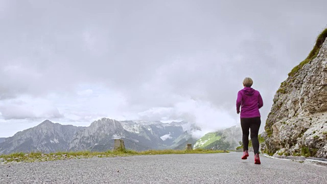 一个多云的日子里，一名老妇人在高山上的一条路上奔跑视频素材