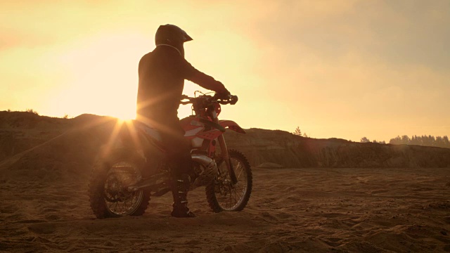 专业FMX摩托车骑手在他的摩托车上休息，俯瞰坚硬的沙地越野地形。太阳设置。视频素材