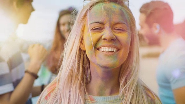 一个美丽的金发女孩在胡里节与她的朋友跳舞的特写肖像。她的脸上和衣服上都涂满了彩色的粉末。视频素材