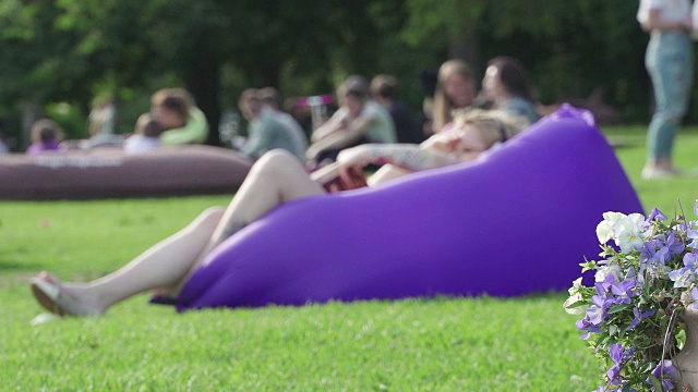 年轻的金发女孩在温暖的阳光明媚的一天躺在一个充气床垫上在绿色的草地上野餐。手里拿着一本书，读着。其余的学生或年轻人在城市公园里。健康的生活方式和娱乐的概念视频下载