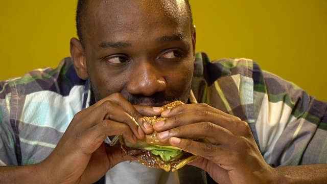 非裔美国人与一个有趣的面部表情吃一个汉堡在一个咖啡馆的特写慢动作视频下载