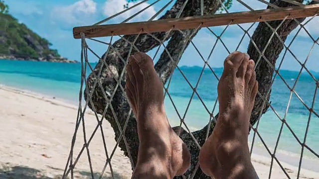 在白色的沙滩和蓝色的海洋前，赤裸的男人的脚在吊床上晃荡视频素材