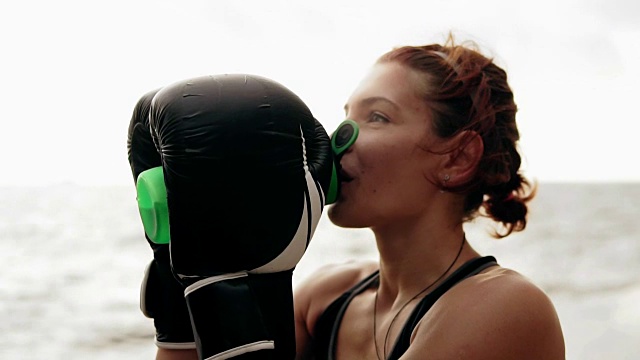 口渴的女拳击手戴着手套，在训练后休息时饮用水瓶里的水。在海边训练的美女视频下载