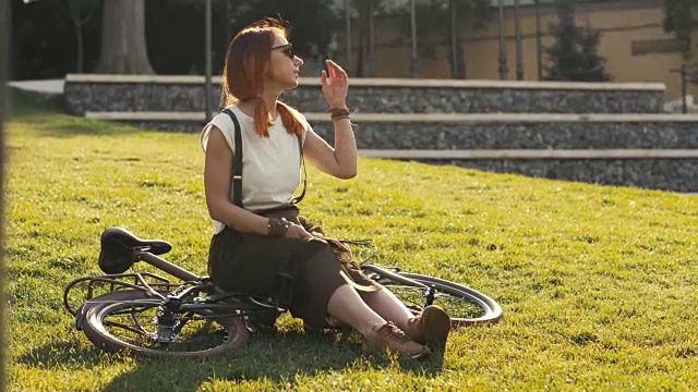 红头发的女人坐在自行车上躺在城市公园的草地上。女子自行车公园视频素材