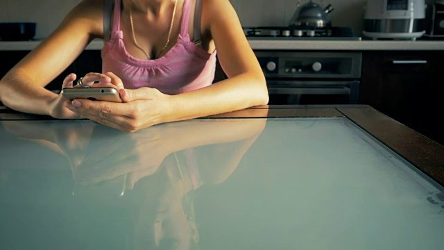 多莉，这个年轻的女孩坐在家里厨房的桌子上，在智能手机上阅读。视频素材