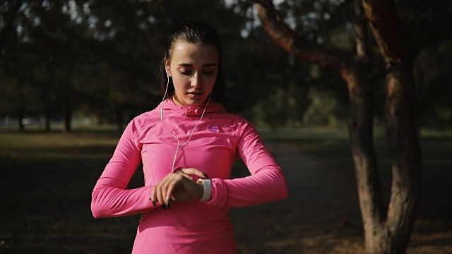 漂亮的女运动员穿着粉红色运动服在户外慢跑，健康的生活方式理念。缓慢的运动。她看到了秒表上的时间。视频素材