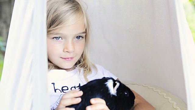 金发小女孩坐在或躺在吊床上，抱着她的宠物豚鼠。视频素材