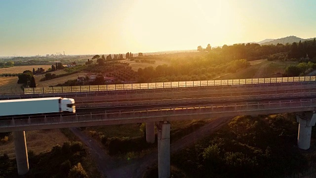 空中拍摄的一个白色卡车与货物装载移动在巨大的桥上与交通在农村地区。背景是美丽的自然风景。视频素材