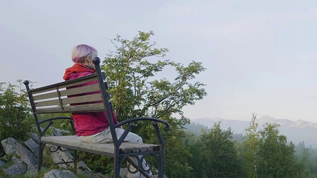 孤独年轻的金发女人坐在公园长椅上的背影。女孩独自坐在长椅上看自然景观视频下载