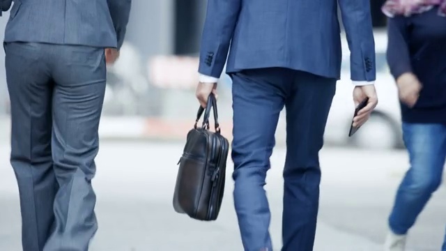 男性和女性商务人士走在繁忙的大城市街道上的后视图。他们都穿着时尚的灰/蓝西装，男人拿着包。他们急着去参加投资者会议。视频素材