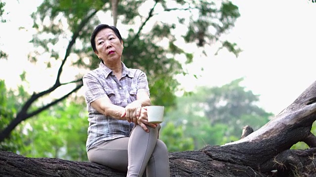 孤独的亚洲老年妇女坐在公园里独自喝咖啡，看起来很悲伤，思考着生活视频素材