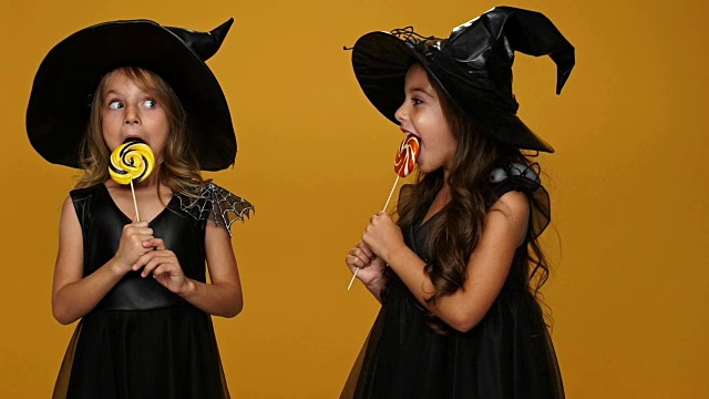 漂亮的小女孩戴着装饰好的裙子和帽子吃着棒棒糖孤立在橙色上视频下载