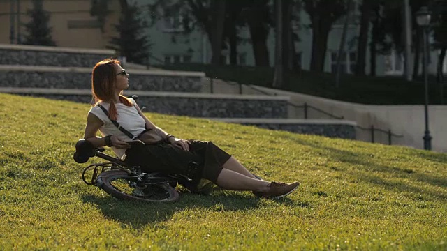 红头发的女人骑自行车躺在城市公园的绿色草地上。女子自行车公园视频素材