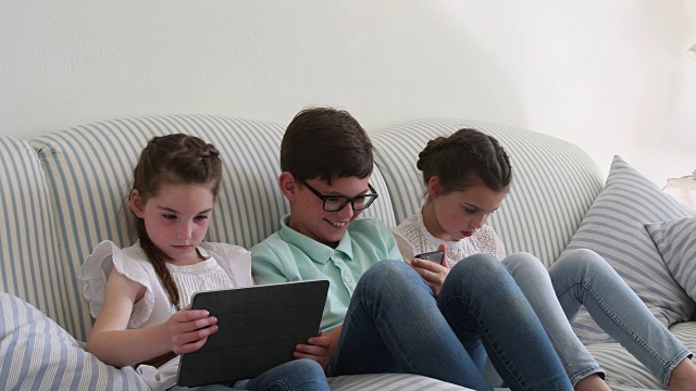 两个女孩和他们的兄弟在沙发上使用平板电脑和手机视频素材