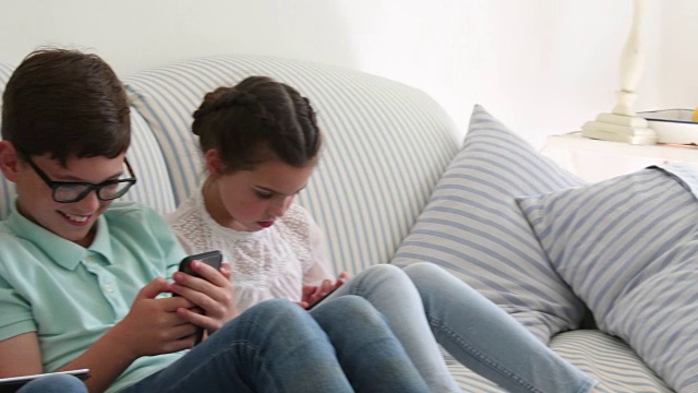 两姐妹和兄弟在家里的沙发上使用数码设备视频素材