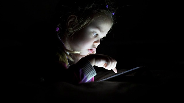 美丽的小女孩看着平板躺在床上的黑暗视频素材