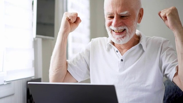 英俊的老年人在家里用笔记本电脑工作。收到好消息又兴奋又高兴视频素材