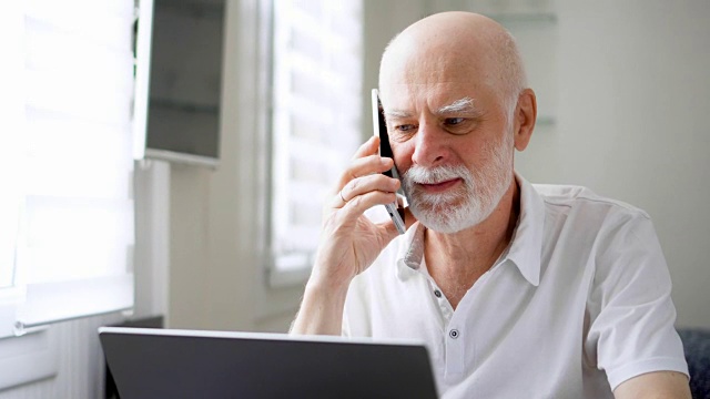 老男人坐在家里拿着笔记本电脑和智能手机。用手机在屏幕上讨论项目视频素材