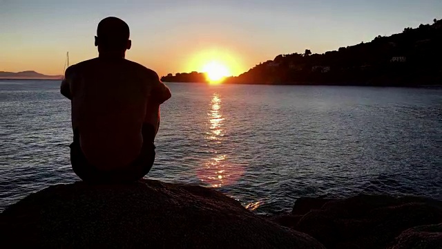 欣赏日落。一个男人坐在岩石沙滩上看风景视频下载