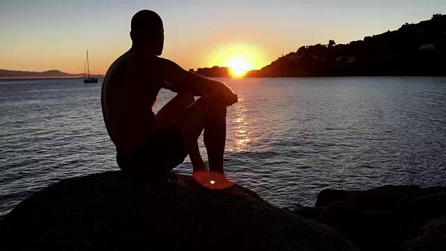 欣赏日落。一个男人坐在岩石沙滩上看风景视频下载