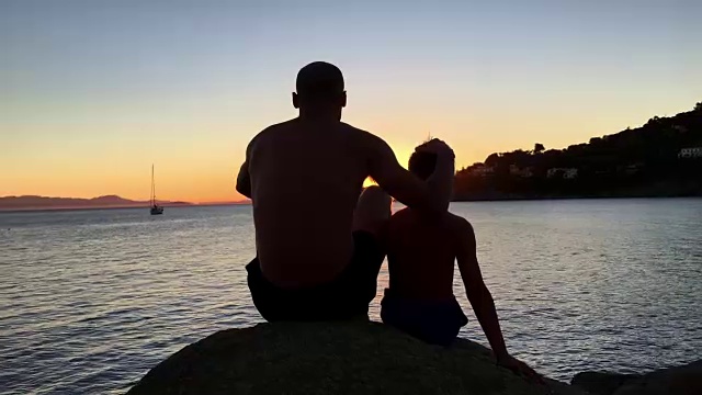 欣赏日落。父亲和儿子坐在岩石海滩上看风景视频下载