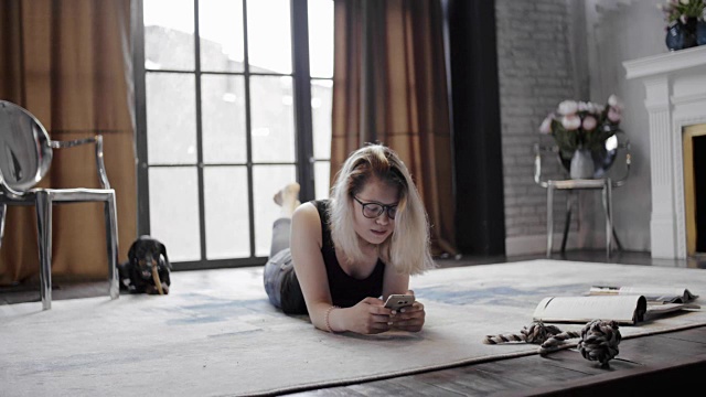 女孩戴着眼镜拿着手机躺在地板上，背景是腊肠犬视频素材