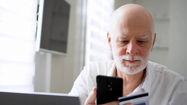 留胡子的帅老头坐在家里。在智能手机上用信用卡网上购物视频素材