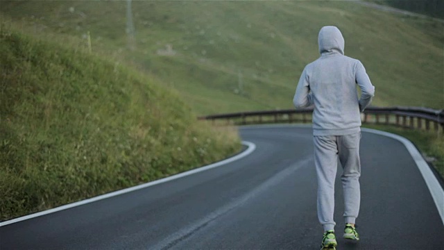 人在乡村山路上慢跑，在大自然中近观慢动作。戴着头巾的男性身影在当地柏油路的清晨斜坡背景下跑步，穿着灰色运动服和运动鞋视频素材