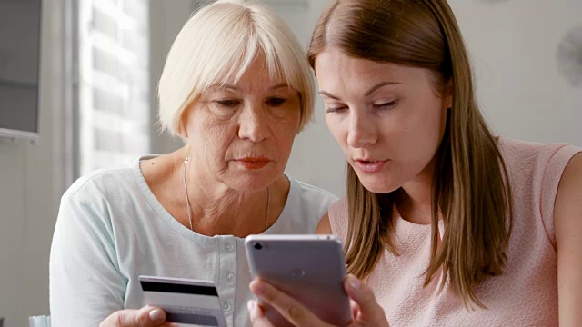 年长的母亲和她的小女儿在家里使用智能手机。用手机刷卡购物视频素材
