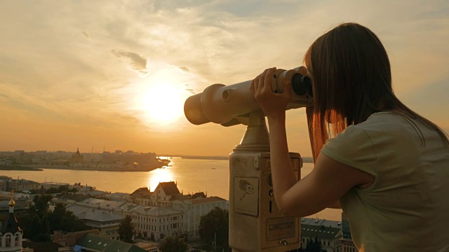 年轻女子透过旅游望远镜，在夕阳下探索城市视频下载